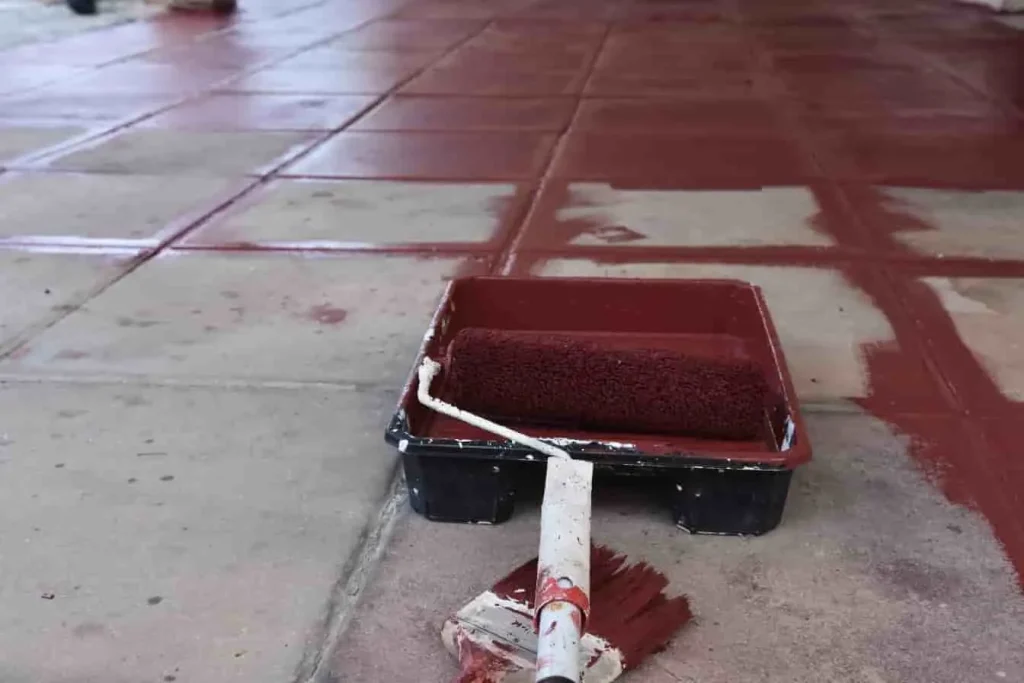 Peinture brun-rouge appliquée sur sol en béton carrelé avec rouleau, transition visible entre zones peintes et brutes