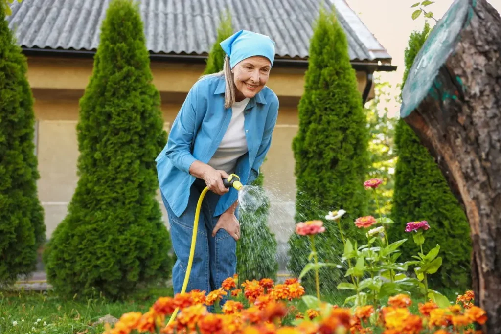 Une femme souriante arrose des fleurs orange et roses pour attirer les papillons dans le jardin tout en prenant soin de la biodiversité de son espace vert
