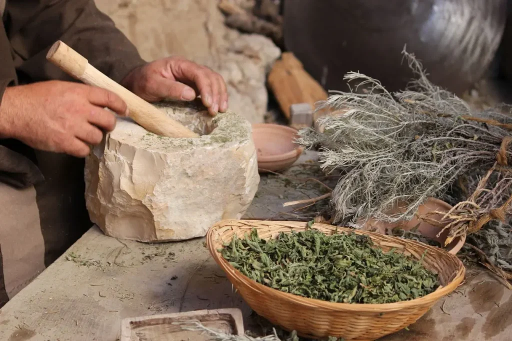 Personne utilisant pilon en bois dans mortier de pierre avec feuilles séchées, illustrant la méthode artisanale pour sécher les herbes aromatiques