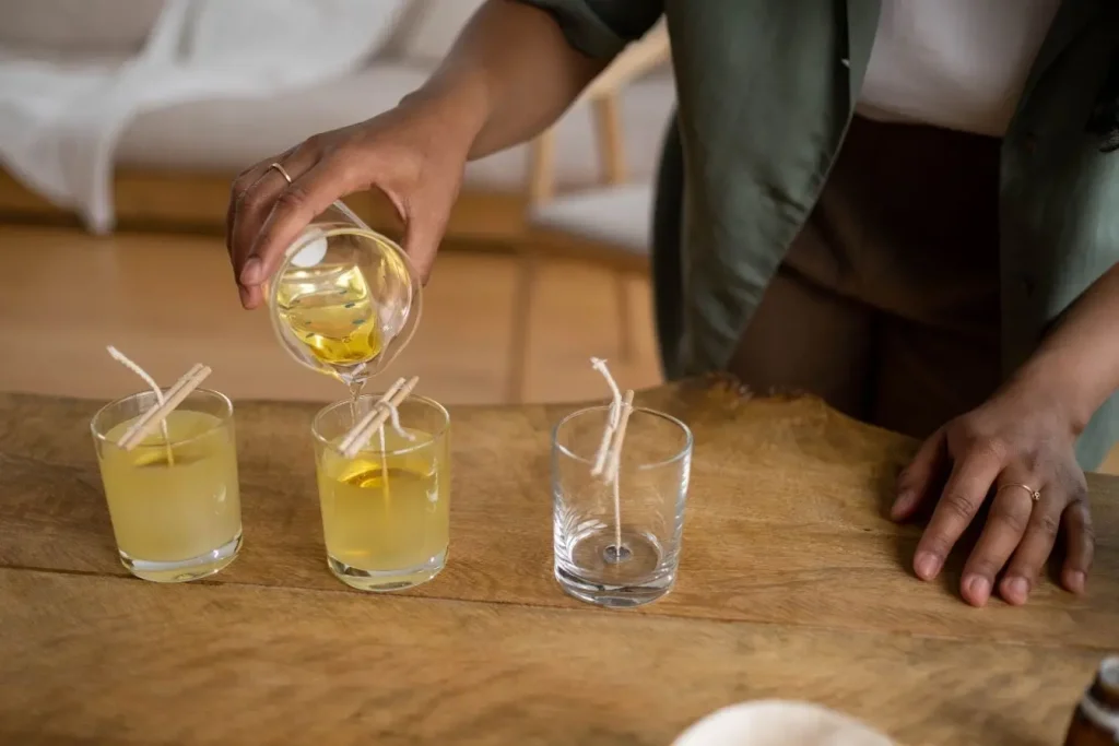 Personne verse cire fondue dans bocal en verre avec mèche maintenue, image illustrant la fabrication artisanale de bougies naturelles.