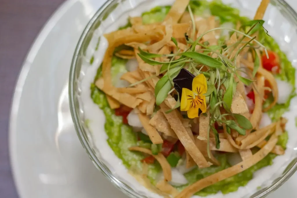 Bol en verre garni de purée verte et crème blanche, décoré de légumes frais, tortillas croustillantes et fleur jaune violette comestible.
