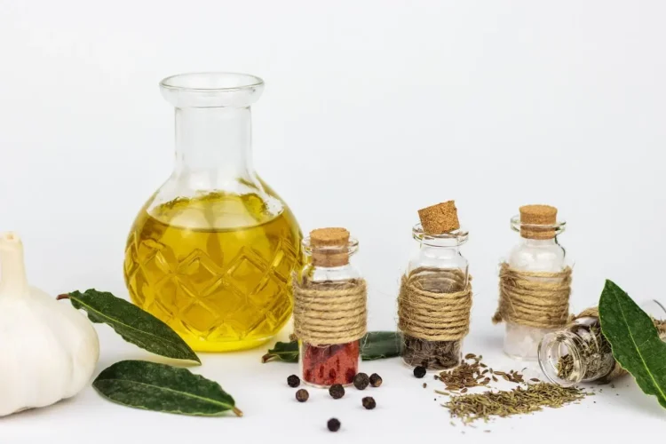 Bouteille d’huile jaune posée sur table blanche entourée de pots d’épices et feuilles de laurier, image illustrant une huile infusée aux herbes aromatiques.