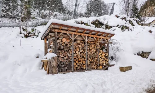 Abri en bois rempli de bûches empilées sous toit enneigé, image qui illustre la préparation d’un abri à bois de chauffage en hiver.