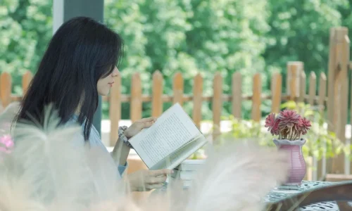 Personne assise à une table en bois lisant un livre, entourée de verdure et fleurs, image qui illustre un coin lecture paisible au jardin.