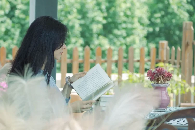 Personne assise à une table en bois lisant un livre, entourée de verdure et fleurs, image qui illustre un coin lecture paisible au jardin.