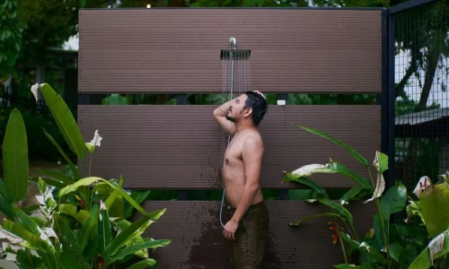 Homme en short sous eau claire avec plantes tropicales, image représentative d’une douche de jardin.