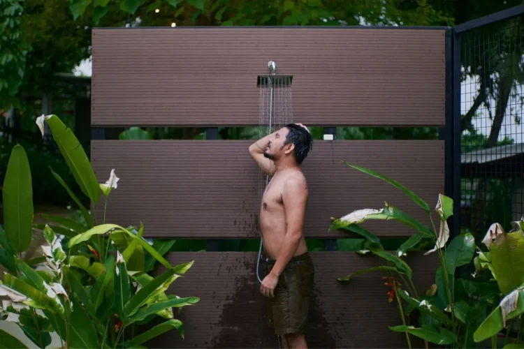 Homme en short sous eau claire avec plantes tropicales, image représentative d’une douche de jardin.