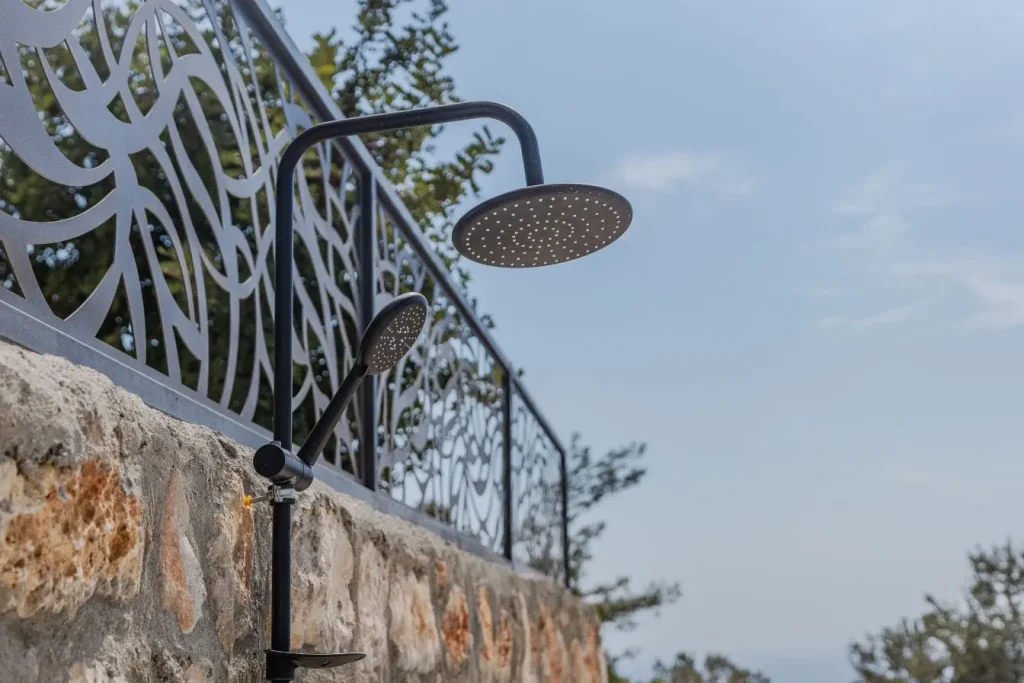 Douche extérieure fixée sur mur en pierre avec pommeau rond et ciel bleu, reflet d’un aménagement élégant de douche de jardin.