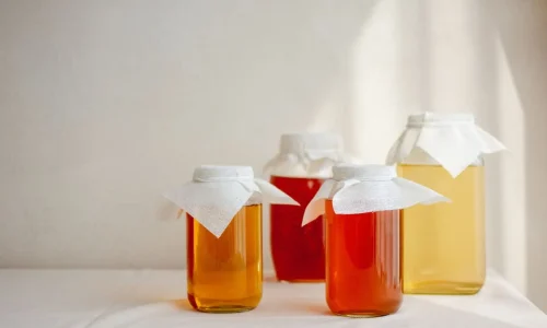 Quatre bocaux en verre remplis de liquides ambrés recouverts de tissus blancs, image qui illustre la préparation artisanale du kombucha maison.