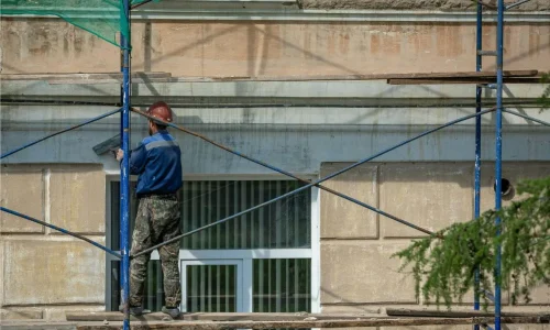 Ouvrier en casque rouge appliquant enduit sur mur beige depuis échafaudage, reflet d’un ravalement de façade réalisé soi‑même.