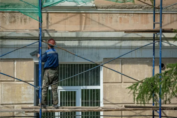 Ouvrier en casque rouge appliquant enduit sur mur beige depuis échafaudage, reflet d’un ravalement de façade réalisé soi‑même.
