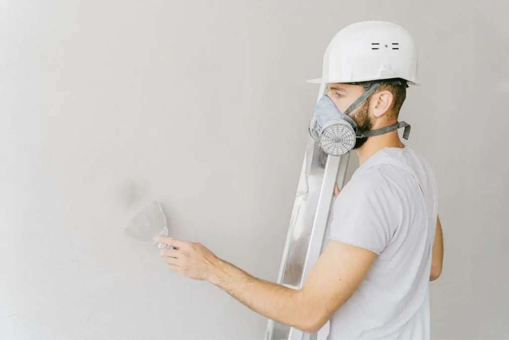 Personne en casque blanc et masque respiratoire grattant mur blanc avec outil, reflet d’un ravalement de façade réalisé soi‑même.