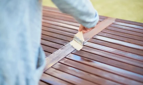 Gros plan sur main appliquant vernis au pinceau sur lames de bois, image qui illustre l’entretien d’une terrasse en bois exotique.
