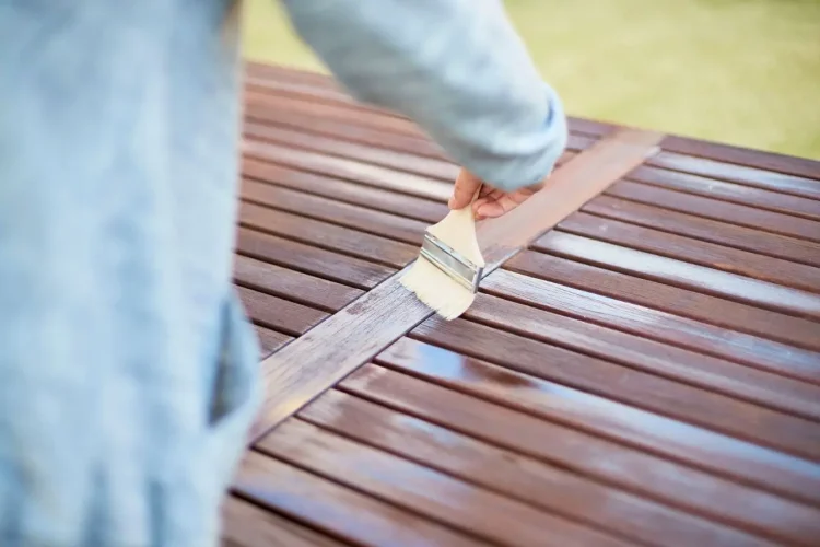 Gros plan sur main appliquant vernis au pinceau sur lames de bois, image qui illustre l’entretien d’une terrasse en bois exotique.