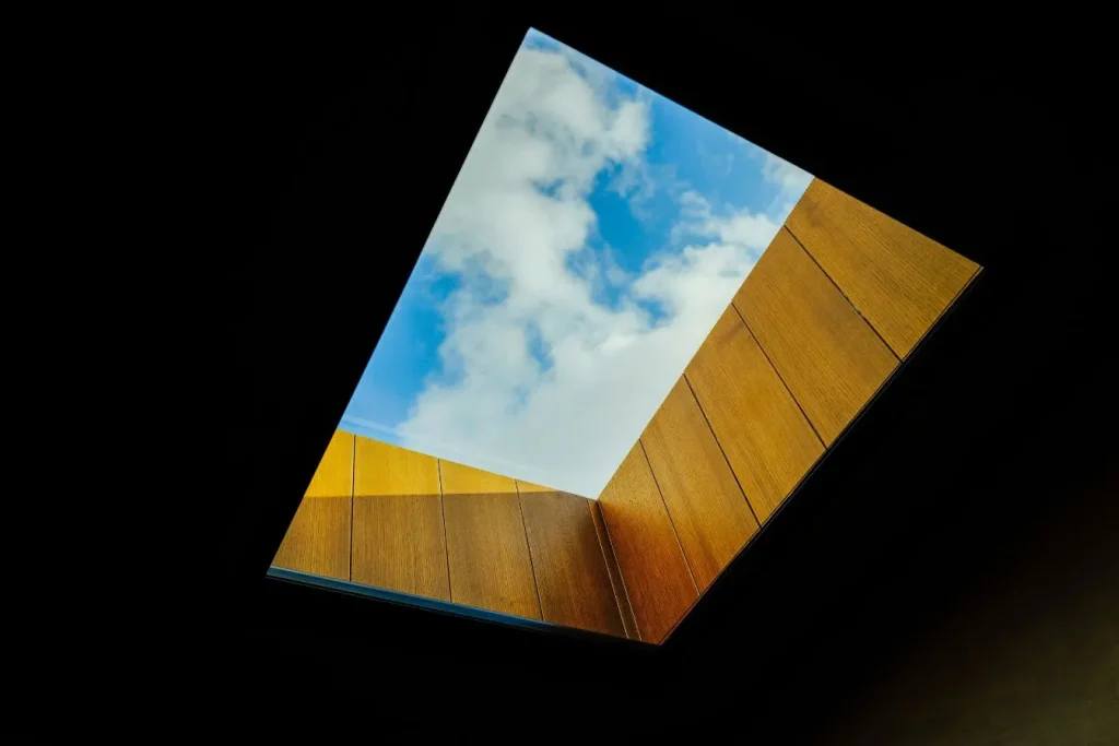 Vue intérieure d’un plafond en bois avec ouverture rectangulaire laissant entrer ciel bleu et nuages, reflet d’un puits de lumière naturel.