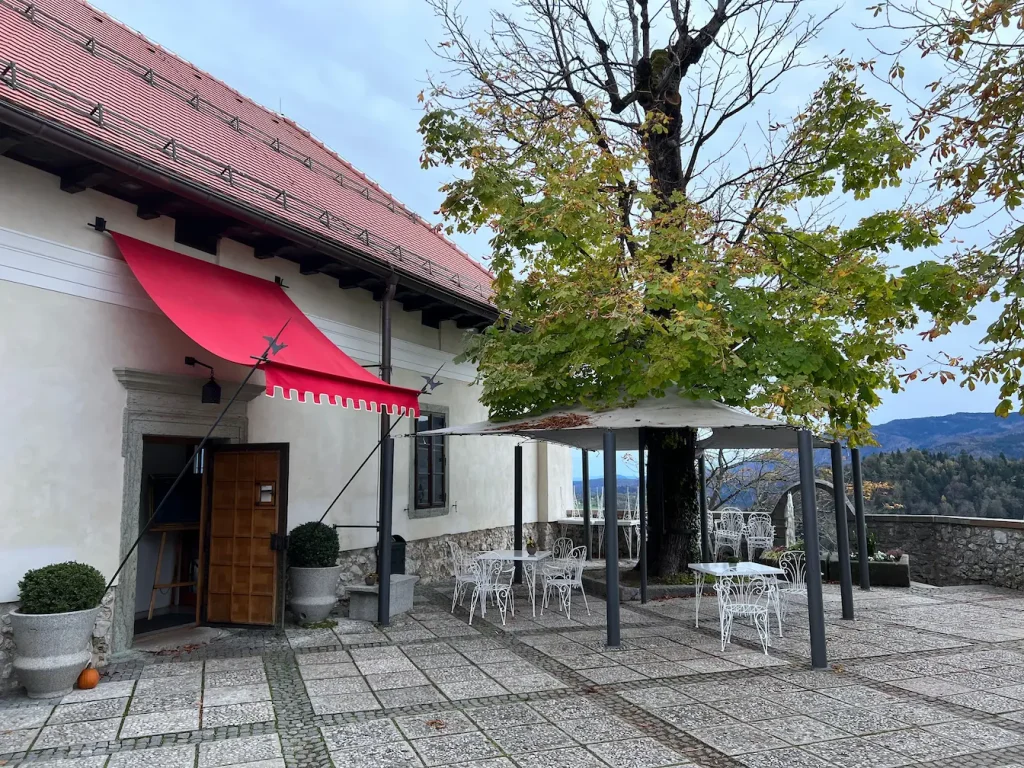 Terrasse pavée avec tables blanches sous grand arbre, protégée par auvent rouge fixé au mur, symbole d’une installation réussie.